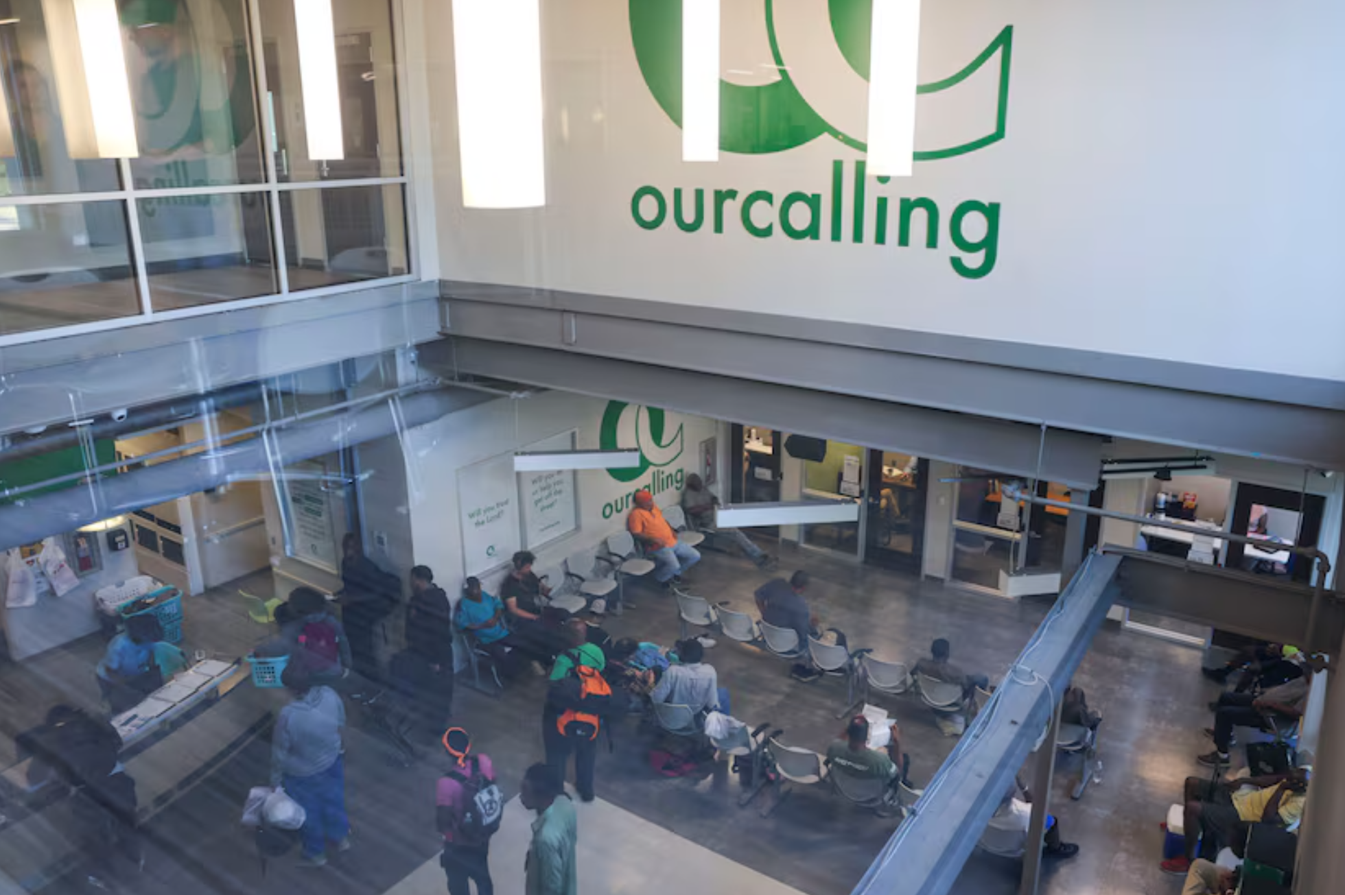 Indoor view of a community center lobby with “ourcalling” logo on the wall. Several people are seated in rows of chairs, while others stand near counters and tables. Bright overhead lights and glass partitions are visible.