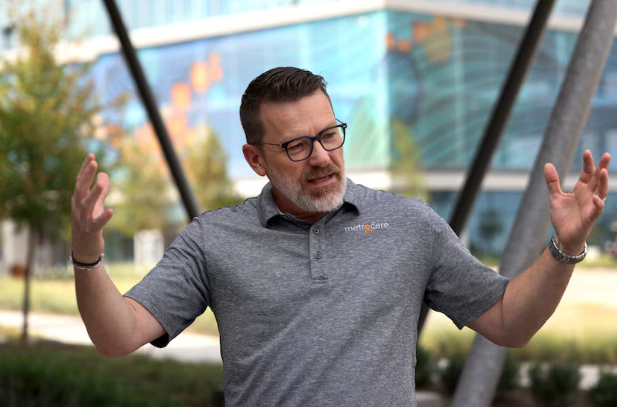 Person wearing a gray polo shirt with the “MetroCare” logo, standing outdoors in front of a modern glass building, gesturing with both hands raised.