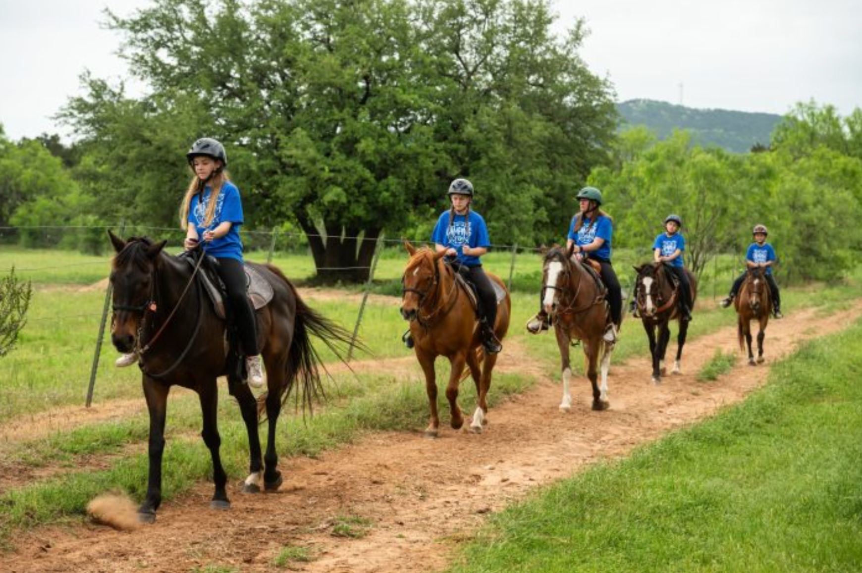 Group of riders in blue shirts on horseback following a dirt trail through a green field with trees in the background.