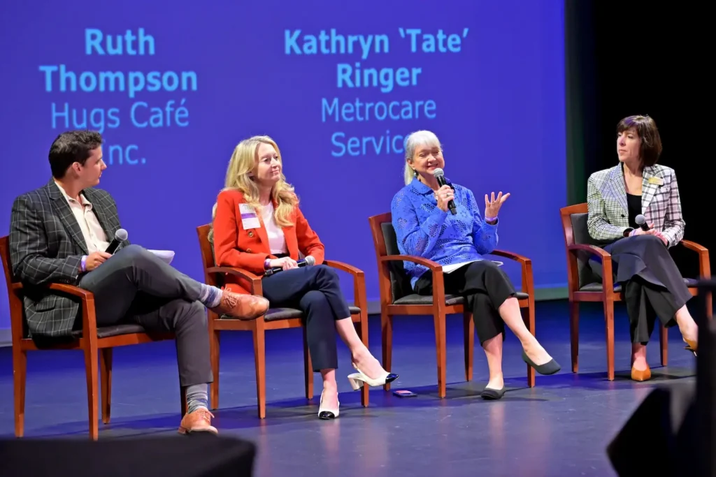 Panel discussion on a stage with four seated speakers holding microphones. Names and affiliations are projected behind them on a blue backdrop, including “Ruth Thompson, Hugs Café Inc.” and “Kathryn ‘Tate’ Ringer, Metrocare Services.”