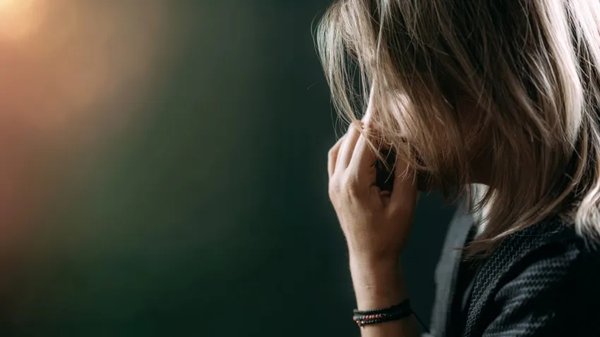 Side-profile close-up of a person with shoulder-length hair, hand touching their face, wearing a dark top and bracelet, against a softly blurred background with warm-to-dark gradient lighting.