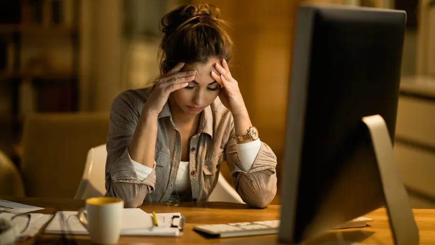 In The News 3 Person sitting at a desk in front of a computer monitor, holding both temples with hands while looking downward. The desk contains a keyboard, papers, a smartphone, a pen, and a mug. The scene is warmly lit, with blurred shelves or furniture in the background.