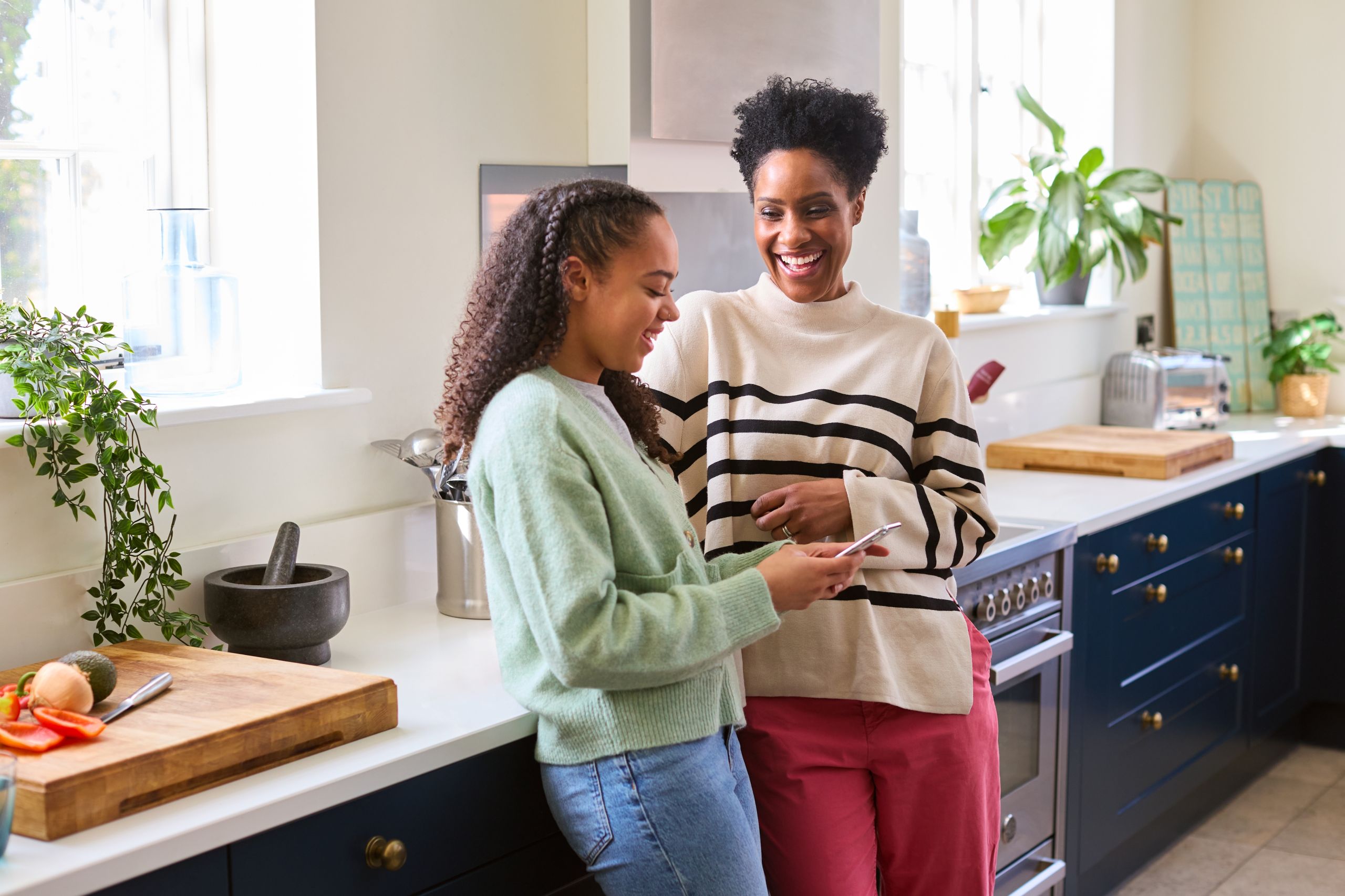 Two people stand together in a bright, modern kitchen, looking at a smartphone. One person leans against a white countertop while holding the phone, and the other stands nearby. The kitchen features dark blue cabinets, white countertops, potted plants, a cutting board with vegetables, and natural light streaming through large windows.